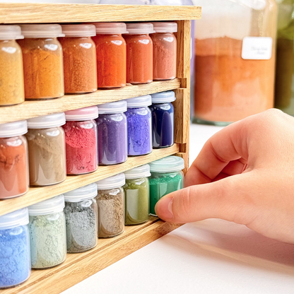 Colorful powders in small jars on a wooden shelf with a hand reaching out.