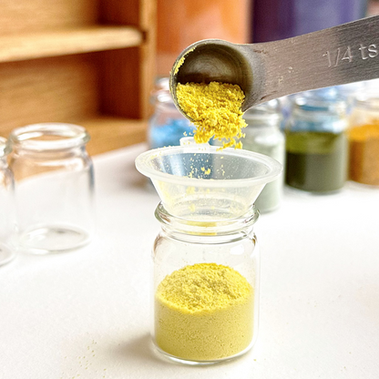 Yellow weld pigment being poured from a measuring spoon into a glass container on a kitchen counter.