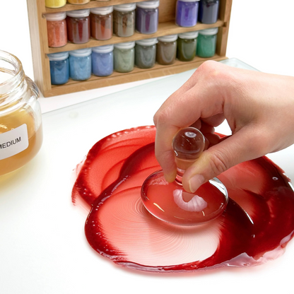 Hand using a small container to mix red paint on a white surface with jars of paint supplies in the background.