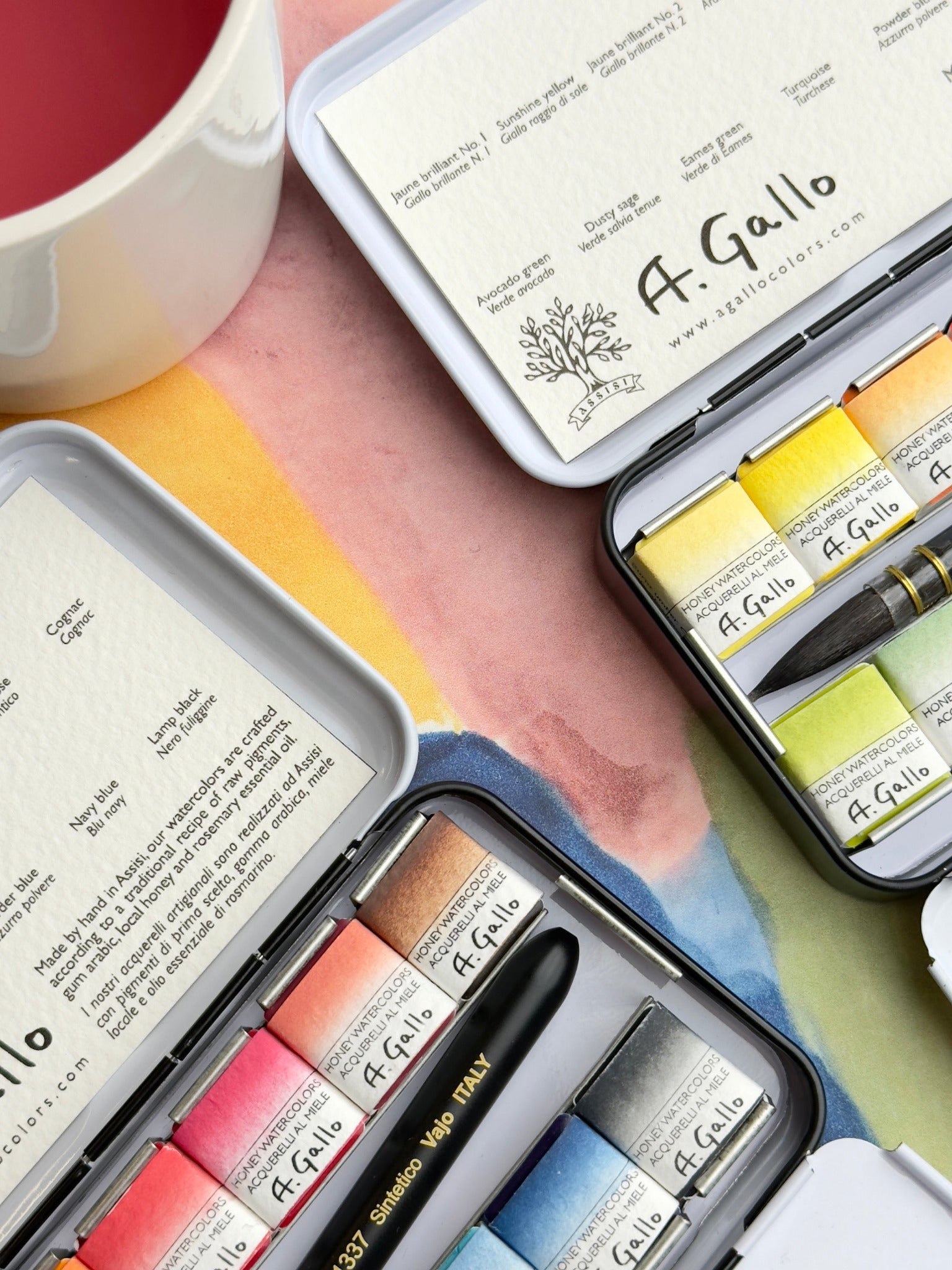 A. Gallo watercolor paint set with a cup of tea and a book on a colorful surface.