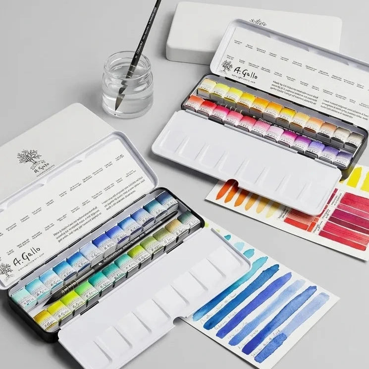 An open set of watercolor paints in a white metal case, shown alongside closed cases, swatch cards, a paintbrush, and a small jar of water on a light gray background. The brand name 'L'Enseigne' is visible on the paint tubes.